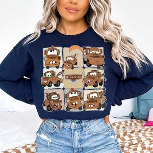 Może przedstawiać: Granatowa bluza z nadrukiem w kratkę z kresk&oacute;wkowymi obrazkami Złomka z filmu Auta. Obrazy są w odcieniach brązu i beżu, ze słowem "Cars" pośrodku. Bluza ma okrągły dekolt.