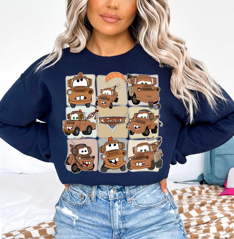 Może przedstawiać: Granatowa bluza z nadrukiem w kratkę z kresk&oacute;wkowymi obrazkami Złomka z filmu Auta. Obrazy są w odcieniach brązu i beżu, ze słowem "Cars" pośrodku. Bluza ma okrągły dekolt.