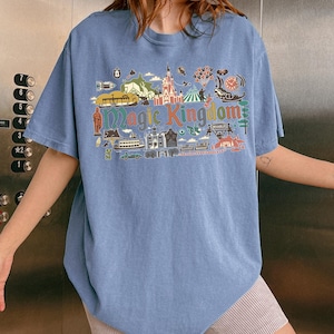 May include: A light blue t-shirt with a colorful graphic that reads "Magic Kingdom." The graphic features various illustrations of buildings, rides, and other elements associated with a theme park. The shirt has short sleeves and a relaxed fit.
