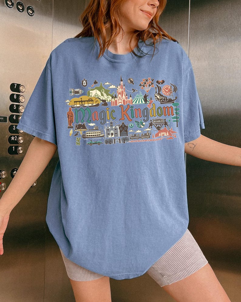 May include: A light blue t-shirt with a colorful graphic that reads "Magic Kingdom." The graphic features various illustrations of buildings, rides, and other elements associated with a theme park. The shirt has short sleeves and a relaxed fit.