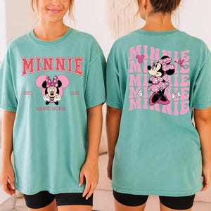 May include: A teal t-shirt featuring Minnie Mouse. The front of the shirt has the word "MINNIE" in pink block letters above a graphic of Minnie Mouse. The back of the shirt has a repeating pink graphic of Minnie Mouse with the word "MINNIE".