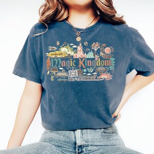 May include: A navy blue t-shirt featuring a colorful graphic of the Magic Kingdom. The design includes illustrations of a castle, a carousel, and other park icons. The words "Magic Kingdom" are prominently displayed.