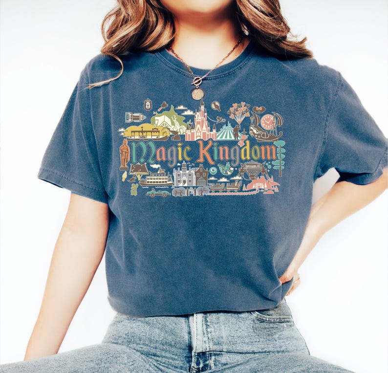 May include: A navy blue t-shirt featuring a colorful graphic of the Magic Kingdom. The design includes illustrations of a castle, a carousel, and other park icons. The words "Magic Kingdom" are prominently displayed.