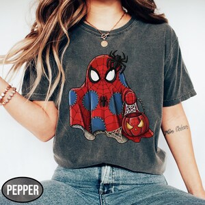 Op de afbeelding: Donkergrijs T-shirt met een Halloween-thema. De afbeelding toont een geest in een Spider-Man-kostuum die een pompoenvormige snoepemmer vasthoudt. Een zwarte spin zit op het hoofd van de geest. Het woord "PEPPER" staat linksonder.