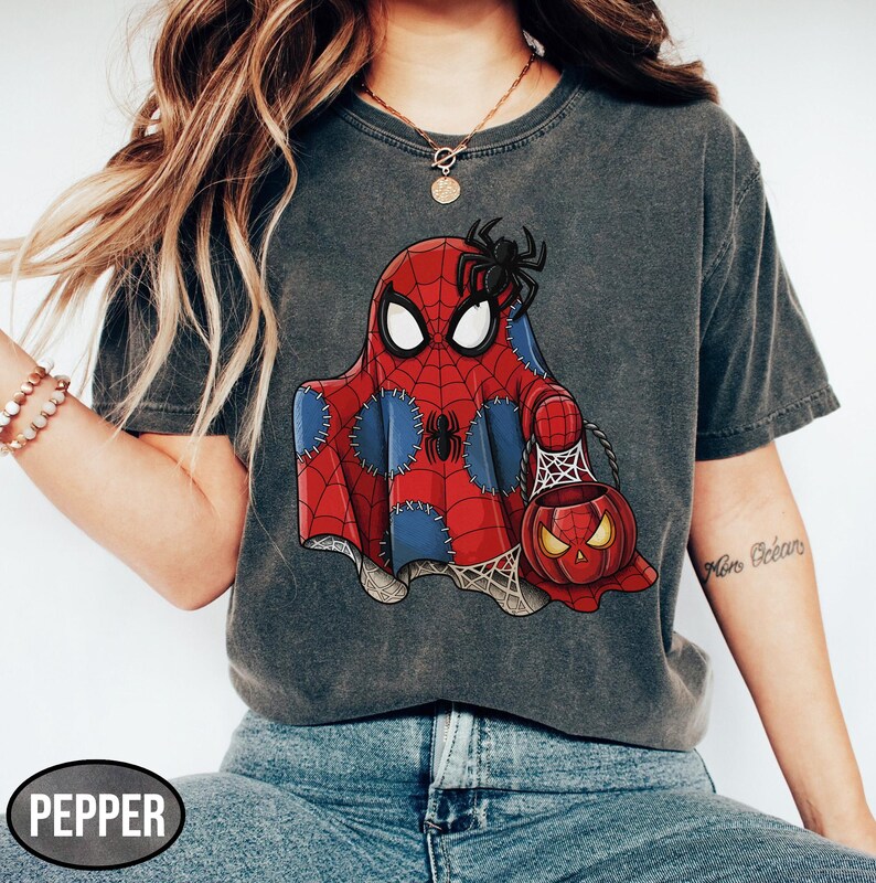 Op de afbeelding: Donkergrijs T-shirt met een Halloween-thema. De afbeelding toont een geest in een Spider-Man-kostuum die een pompoenvormige snoepemmer vasthoudt. Een zwarte spin zit op het hoofd van de geest. Het woord "PEPPER" staat linksonder.