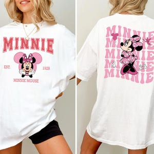 May include: White t-shirt featuring Minnie Mouse. The front design includes the word "MINNIE" in pink, with "EST. 1928" and "MINNIE MOUSE" below. The back displays a pink Minnie Mouse graphic with the word "MINNIE" repeated in a wavy pattern.