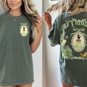 May include: Olive green t-shirt featuring a front and back design. The front has a cameo of a princess in a green dress. The back displays the word "Tiana" and other characters from the movie "Princess and the Frog".