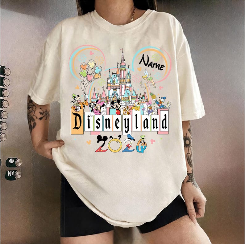 Op de afbeelding: Cr&egrave;mekleurig T-shirt met een kleurrijke Disneyland-afbeelding. Het ontwerp bevat een kasteel, ballonnen en diverse Disney-figuren. De tekst "Disneyland 2020" is prominent aanwezig.