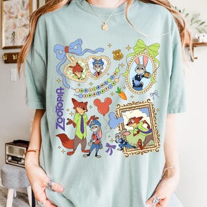 May include: A light green t-shirt featuring colorful cartoon illustrations of characters from the movie "Zootopia." The design includes images of Nick Wilde, Judy Hopps, and other characters, along with the word "Zootopia."