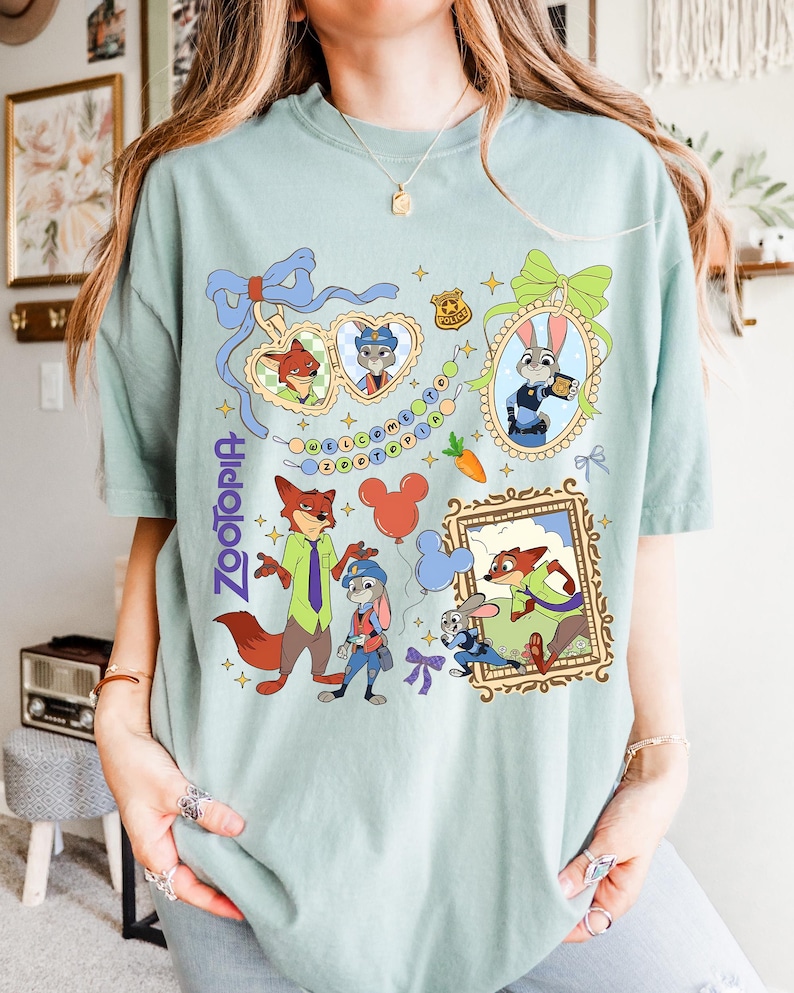 May include: A light green t-shirt featuring colorful cartoon illustrations of characters from the movie "Zootopia." The design includes images of Nick Wilde, Judy Hopps, and other characters, along with the word "Zootopia."