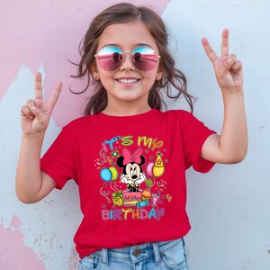 May include: A red t-shirt featuring a graphic of Minnie Mouse with a birthday cake and the text "It's my Birthday". The shirt is worn by a young girl wearing sunglasses and making a peace sign.