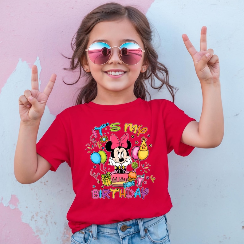May include: A red t-shirt featuring a graphic of Minnie Mouse with a birthday cake and the text "It's my Birthday". The shirt is worn by a young girl wearing sunglasses and making a peace sign.
