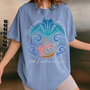May include: A light blue t-shirt featuring a colorful graphic design with the text "There is nowhere you can go that I won't be with you...". The design includes a boat, waves, and a stylized sun.