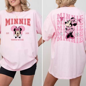 May include: A light pink t-shirt featuring Minnie Mouse. The front of the shirt has the word "MINNIE" in red block letters above a Minnie Mouse graphic. The back of the shirt has a larger Minnie Mouse graphic and the word "MINNIE" repeated.