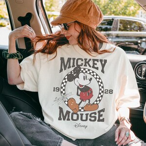 May include: Cream-colored Mickey Mouse t-shirt with a distressed graphic. The design features Mickey Mouse in a circle with a checkered border, the words "Mickey Mouse," and the Disney logo. The shirt has short sleeves.