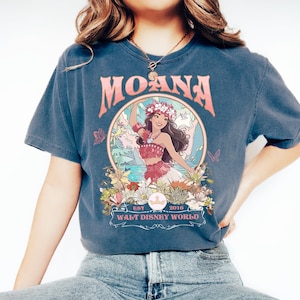 May include: Dark blue t-shirt featuring the word "MOANA" in pink, with a circular graphic of a woman in a floral crown. The graphic includes the text "EST 2016 WALT DISNEY WORLD". The shirt has short sleeves.