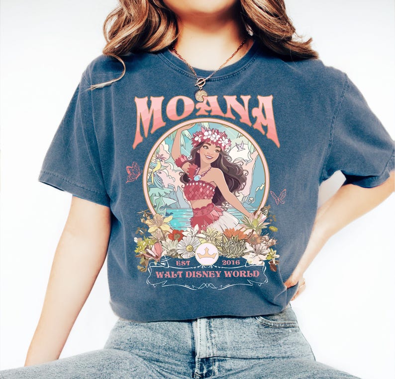 May include: Dark blue t-shirt featuring the word "MOANA" in pink, with a circular graphic of a woman in a floral crown. The graphic includes the text "EST 2016 WALT DISNEY WORLD". The shirt has short sleeves.