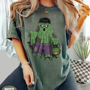 Op de afbeelding: Mosgroen t-shirt met een Halloween-thema. Het ontwerp toont een groene geest met een Frankenstein-achtig uiterlijk, die een pompoen-lantaarnemmer vasthoudt. De geest draagt een paarse broek en heeft zwart haar. Het woord "MOSS" staat linksonder.