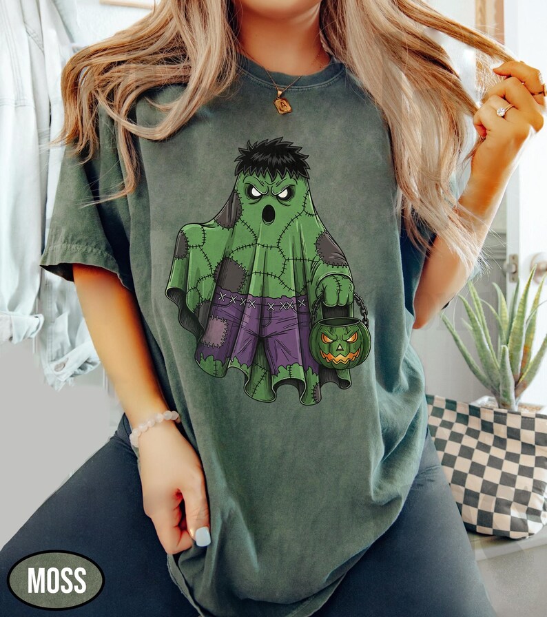 Op de afbeelding: Mosgroen t-shirt met een Halloween-thema. Het ontwerp toont een groene geest met een Frankenstein-achtig uiterlijk, die een pompoen-lantaarnemmer vasthoudt. De geest draagt een paarse broek en heeft zwart haar. Het woord "MOSS" staat linksonder.