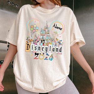 Op de afbeelding: Cr&egrave;mekleurig T-shirt met een kleurrijke Disneyland-afbeelding. Het ontwerp toont een kasteel, Disney-figuren, het woord "Disneyland" en het jaar "2020". Het shirt heeft korte mouwen en een losse pasvorm.