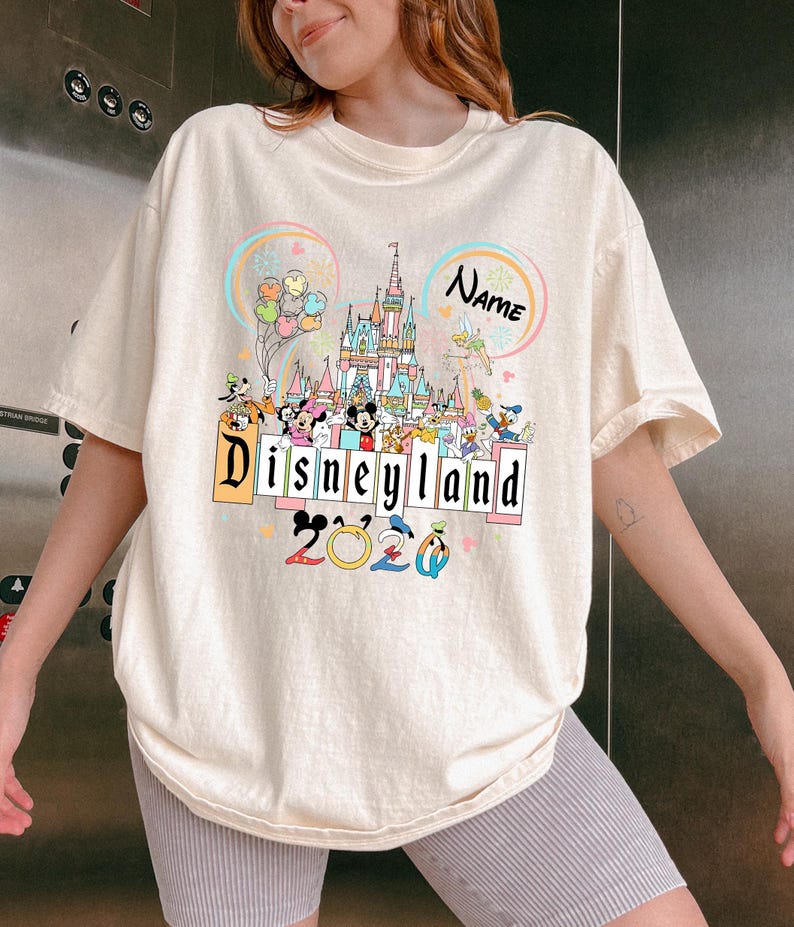 Op de afbeelding: Cr&egrave;mekleurig T-shirt met een kleurrijke Disneyland-afbeelding. Het ontwerp toont een kasteel, Disney-figuren, het woord "Disneyland" en het jaar "2020". Het shirt heeft korte mouwen en een losse pasvorm.