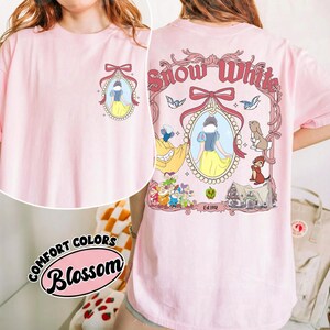 May include: A light pink t-shirt featuring a graphic of Snow White. The front has a small design, while the back showcases a larger illustration with the words "Snow White" and various characters. The shirt is labeled "Comfort Colors Blossom".