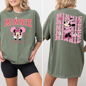 May include: Olive green t-shirt featuring a Minnie Mouse graphic. The front design includes the word "MINNIE" in pink, with a Minnie Mouse illustration below. The back of the shirt has a repeating pink "MINNIE" design with a Minnie Mouse illustration.