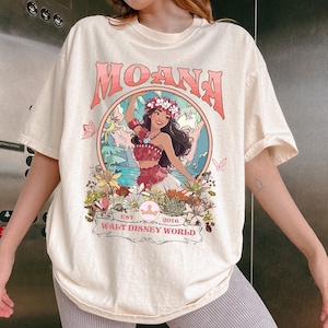 May include: Cream-colored t-shirt featuring a graphic of Moana from Walt Disney World. The design includes the word "MOANA" in pink, a circular illustration of Moana, and the text "EST 2016 WALT DISNEY WORLD".