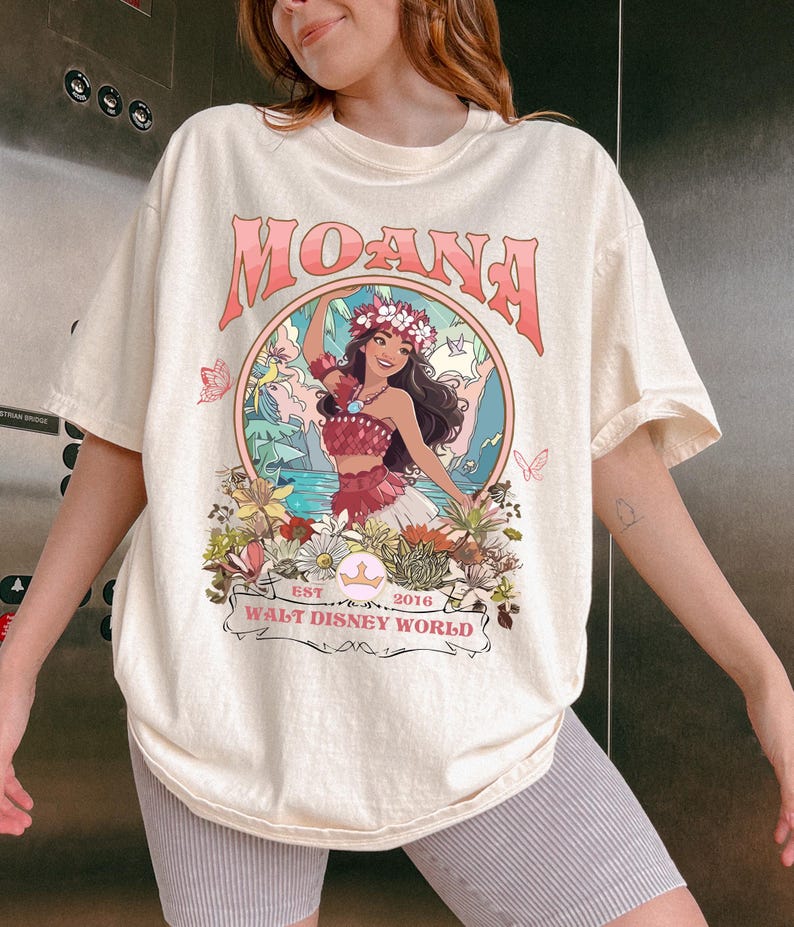 May include: Cream-colored t-shirt featuring a graphic of Moana from Walt Disney World. The design includes the word "MOANA" in pink, a circular illustration of Moana, and the text "EST 2016 WALT DISNEY WORLD".