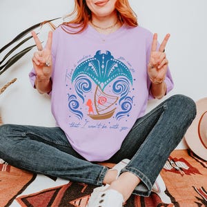 May include: A light purple t-shirt with a graphic of a boat, waves, and a figure. The text on the shirt reads "There is nowhere you can go that I won't be with you." The person is wearing blue jeans.