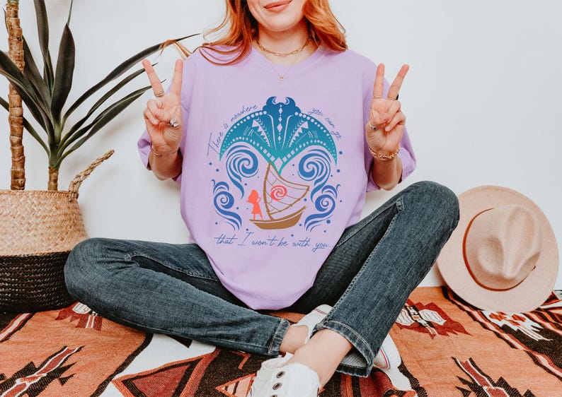 May include: A light purple t-shirt with a graphic of a boat, waves, and a figure. The text on the shirt reads "There is nowhere you can go that I won't be with you." The person is wearing blue jeans.