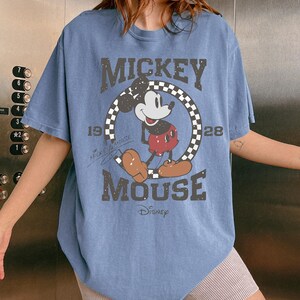 May include: A light blue t-shirt featuring a distressed graphic of Mickey Mouse. The design includes the words "MICKEY MOUSE" and "Disney", along with the year "1928". The shirt has a classic, vintage aesthetic.