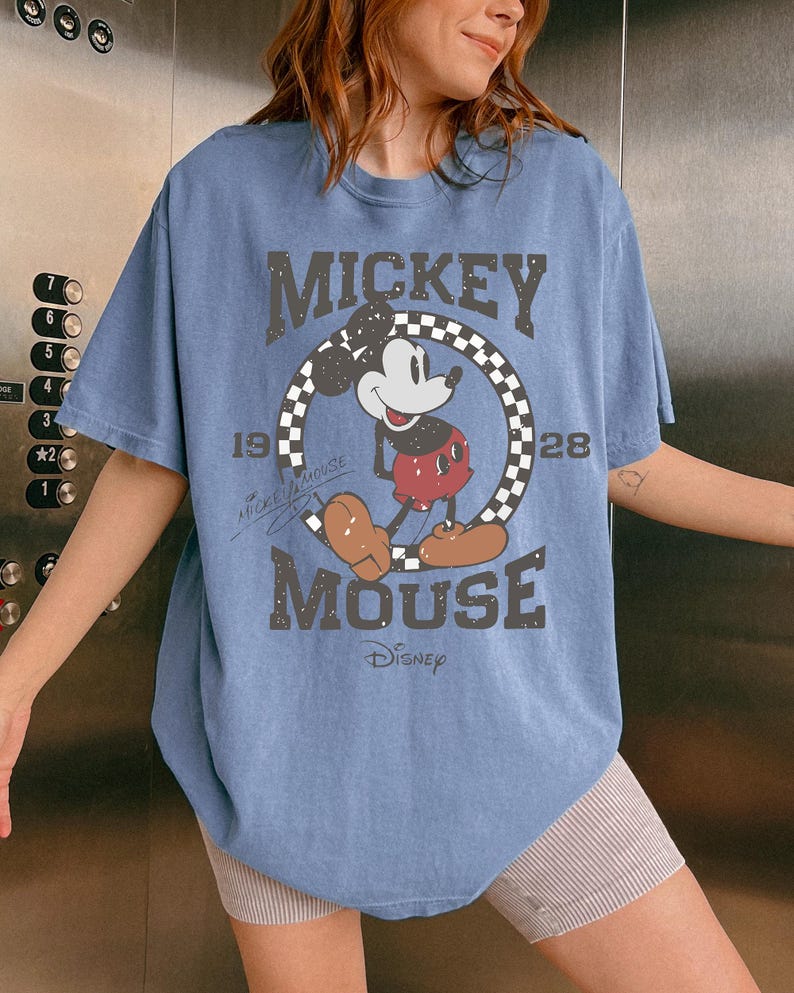 May include: A light blue t-shirt featuring a distressed graphic of Mickey Mouse. The design includes the words "MICKEY MOUSE" and "Disney", along with the year "1928". The shirt has a classic, vintage aesthetic.