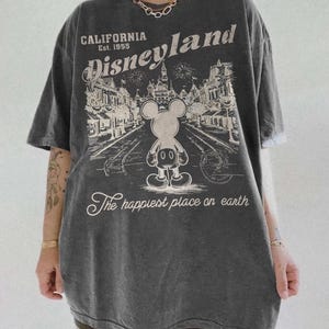 May include: Dark gray oversized t-shirt featuring a Disneyland graphic. The graphic includes the text "California Disneyland Est. 1955" and an illustration of Mickey Mouse. The phrase "The happiest place on earth" is also printed on the shirt.