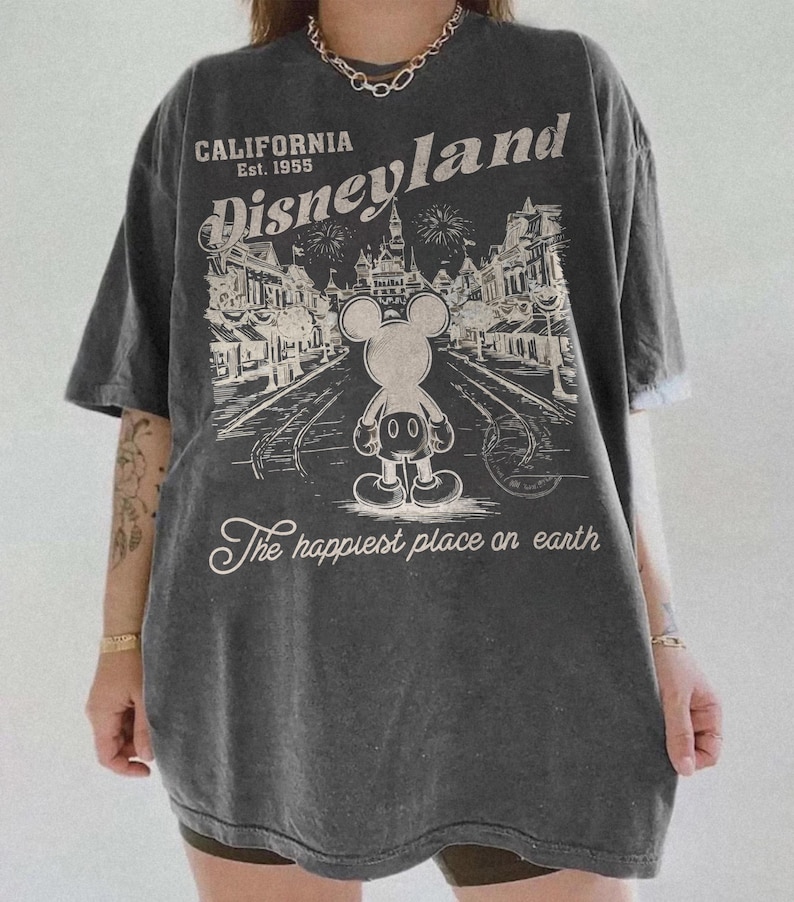May include: Dark gray oversized t-shirt featuring a Disneyland graphic. The graphic includes the text "California Disneyland Est. 1955" and an illustration of Mickey Mouse. The phrase "The happiest place on earth" is also printed on the shirt.