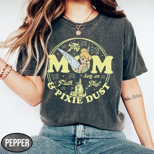 May include: Dark gray t-shirt with a yellow graphic featuring the word "MOM" and Tinkerbell. The design includes the phrases "Faith & Pixie Dust" and "runs on Trust". A castle illustration is also included.
