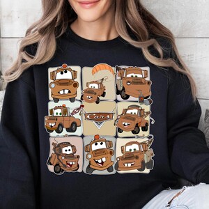 Może przedstawiać: Czarna bluza z nadrukiem w kratkę przedstawiającym postać Złomka z filmu Auta. Obrazy są w odcieniach brązu i beżu, z napisem "Cars" pośrodku.