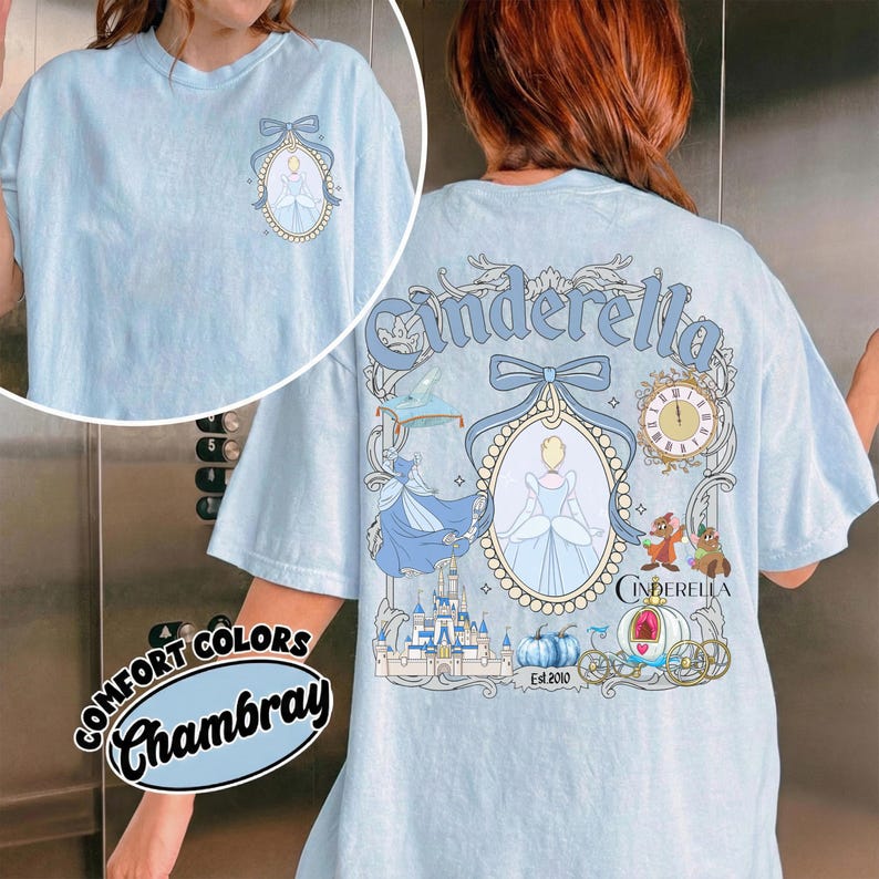 May include: Light blue t-shirt with a Cinderella graphic on the back and a smaller version on the front. The design includes Cinderella, a castle, a pumpkin carriage, and the text "Cinderella". The shirt is made of Comfort Colors Chambray fabric.