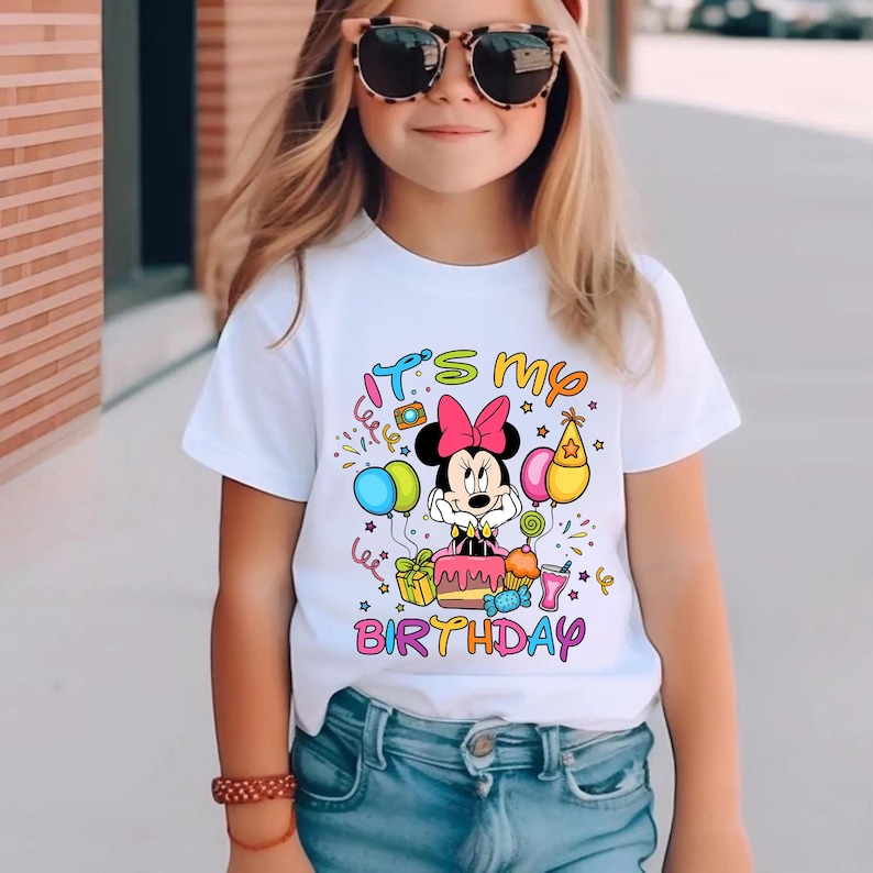 May include: White t-shirt with a colorful graphic featuring Minnie Mouse, balloons, and the text "It's my Birthday." The design includes a pink bow, a birthday cake, and the word "BIRTHDAY" in rainbow colors. The shirt is worn by a young person.