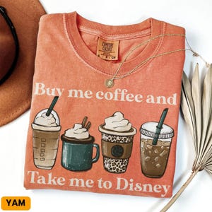 Puede incluir: Camiseta de color coral con el texto "Buy me coffee and Take me to Disney". La camiseta presenta ilustraciones de cuatro bebidas de caf&eacute; diferentes. Un collar de oro est&aacute; drapeado sobre la camiseta.