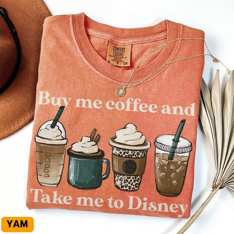 Puede incluir: Camiseta de color coral con el texto "Buy me coffee and Take me to Disney". La camiseta presenta ilustraciones de cuatro bebidas de caf&eacute; diferentes. Un collar de oro est&aacute; drapeado sobre la camiseta.