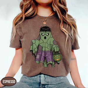 Op de afbeelding: Espresso-kleurig T-shirt met een Halloween-thema. Het ontwerp toont een groene geest met een Frankenstein-achtige uitstraling, een paarse broek en een pompoen-lantaarn. Het shirt is casual kleding.