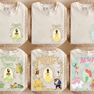 May include: Off-white t-shirts featuring Disney princess designs. The shirts display images of Tiana, Belle, and Ariel, along with other princess-themed graphics. Each shirt has a unique design, with the princess's name and related characters.