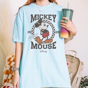 May include: Light blue t-shirt featuring a distressed graphic of Mickey Mouse. The design includes the words "Mickey Mouse" in a vintage font, along with the year "1928" and the Disney logo. The shirt has short sleeves and a relaxed fit.