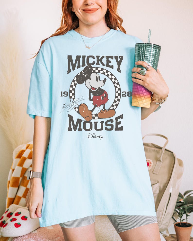 May include: Light blue t-shirt featuring a distressed graphic of Mickey Mouse. The design includes the words "Mickey Mouse" in a vintage font, along with the year "1928" and the Disney logo. The shirt has short sleeves and a relaxed fit.