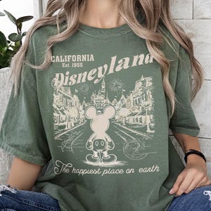 May include: Olive green t-shirt featuring a vintage Disneyland graphic. The design includes the words "California Est. 1955 Disneyland" above an illustration of Mickey Mouse and a park scene. The text "The happiest place on earth" is below the illustration.