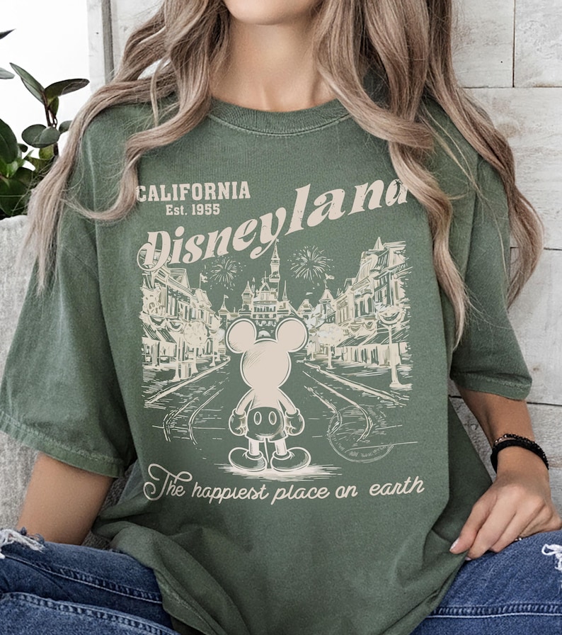 May include: Olive green t-shirt featuring a vintage Disneyland graphic. The design includes the words "California Est. 1955 Disneyland" above an illustration of Mickey Mouse and a park scene. The text "The happiest place on earth" is below the illustration.