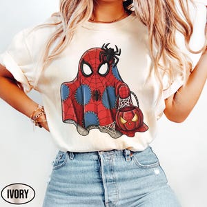 Op de afbeelding: Ivoor kleurig T-shirt met een Halloween-thema ontwerp. De afbeelding toont een spook in een Spider-Man kostuum met een pompoen-vormige snoep emmer. Het ontwerp bevat rood, blauw en zwart, met het woord "IVORY" onderaan.