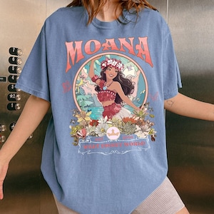 May include: A light blue t-shirt featuring the word "MOANA" in coral lettering above a circular graphic. The graphic depicts a woman in a floral crown and traditional attire, surrounded by flowers and foliage. The text "WALT DISNEY WORLD" is also visible.