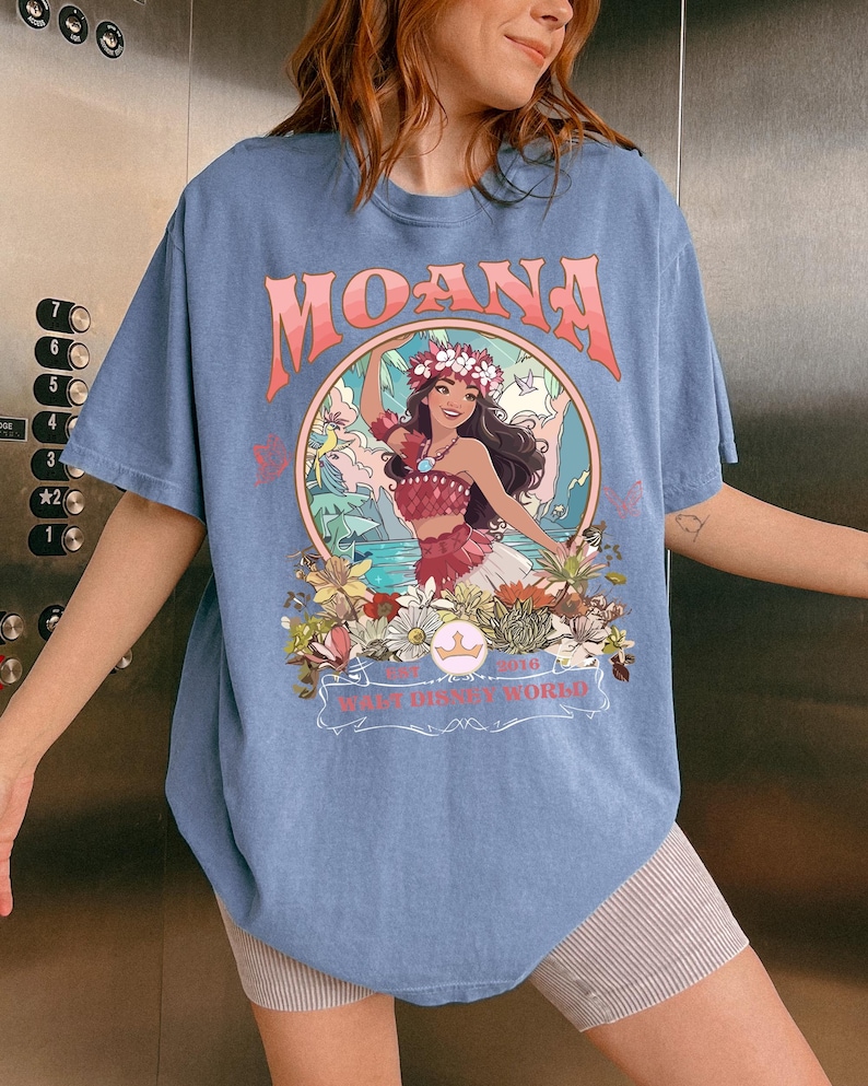 May include: A light blue t-shirt featuring the word "MOANA" in coral lettering above a circular graphic. The graphic depicts a woman in a floral crown and traditional attire, surrounded by flowers and foliage. The text "WALT DISNEY WORLD" is also visible.